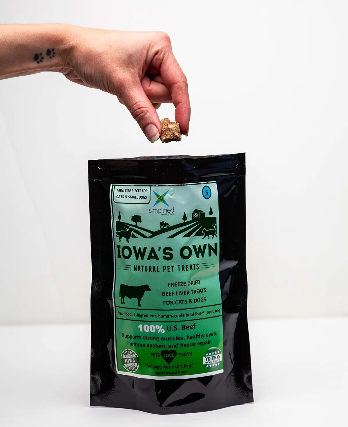 Hand holding a small pet treat above a 'Iowa's Own' pet treats package on a white background